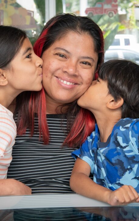 Mother smiling and pictured with two young children kissing her cheeks