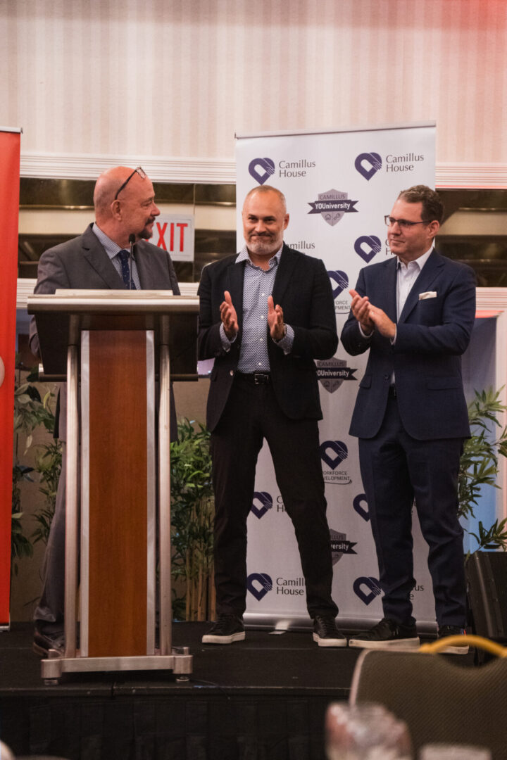 Man giving a speech at a podium while two men applaud behind him